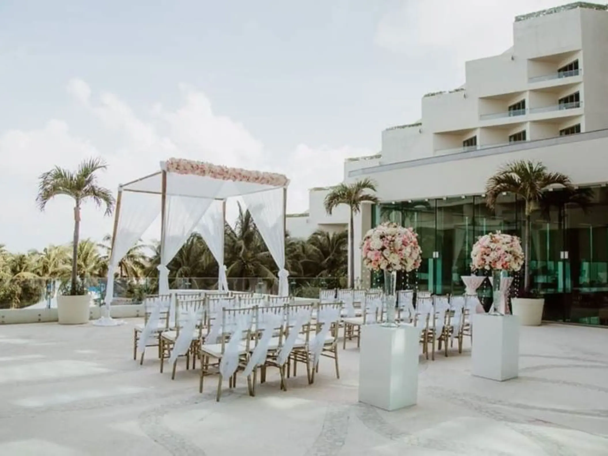 Outdoor wedding venue on a terrace with white chairs, a floral arch, and a view of the resort building.