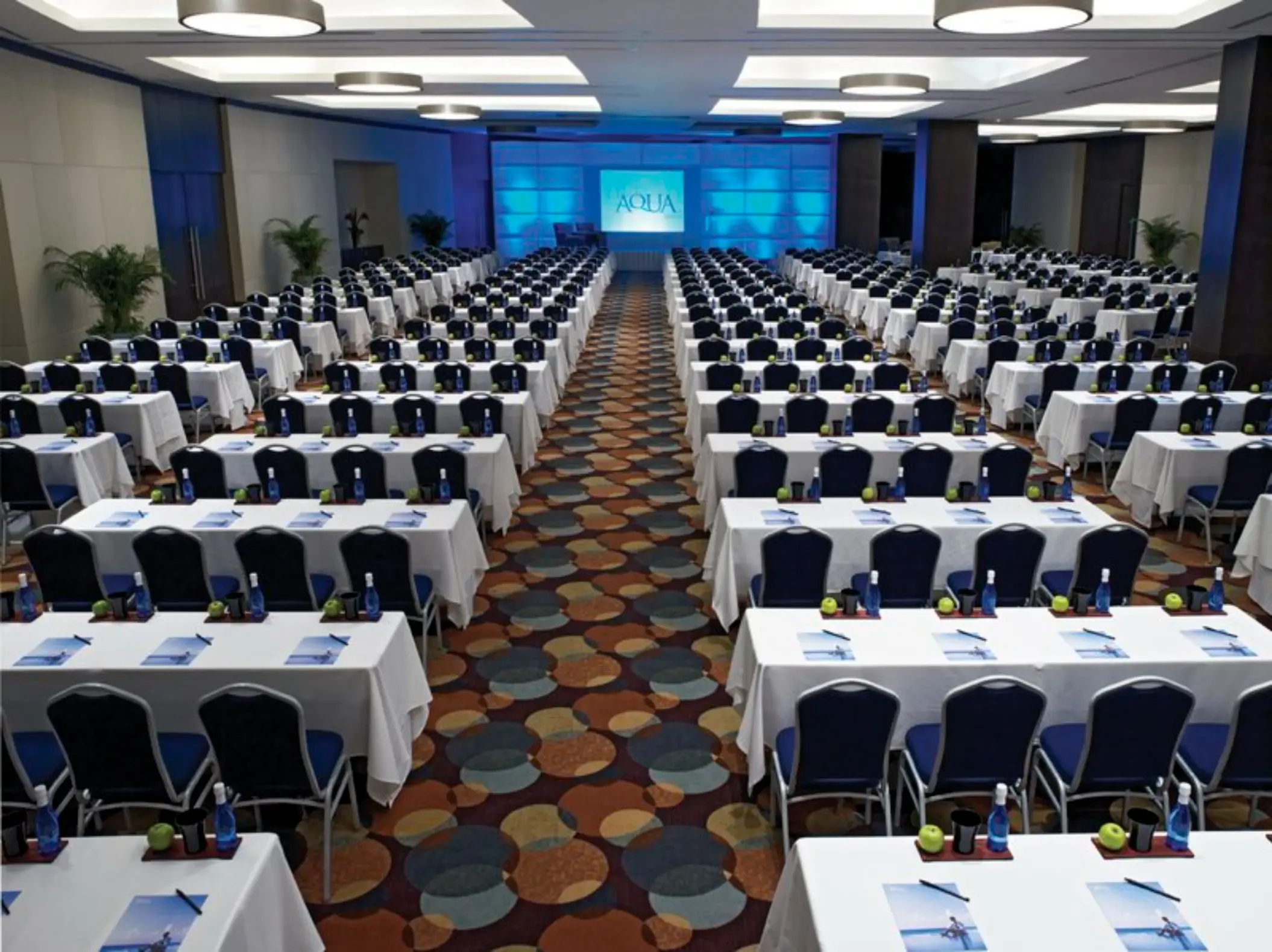 A large conference room set up for a meeting with rows of tables, chairs, and a screen.