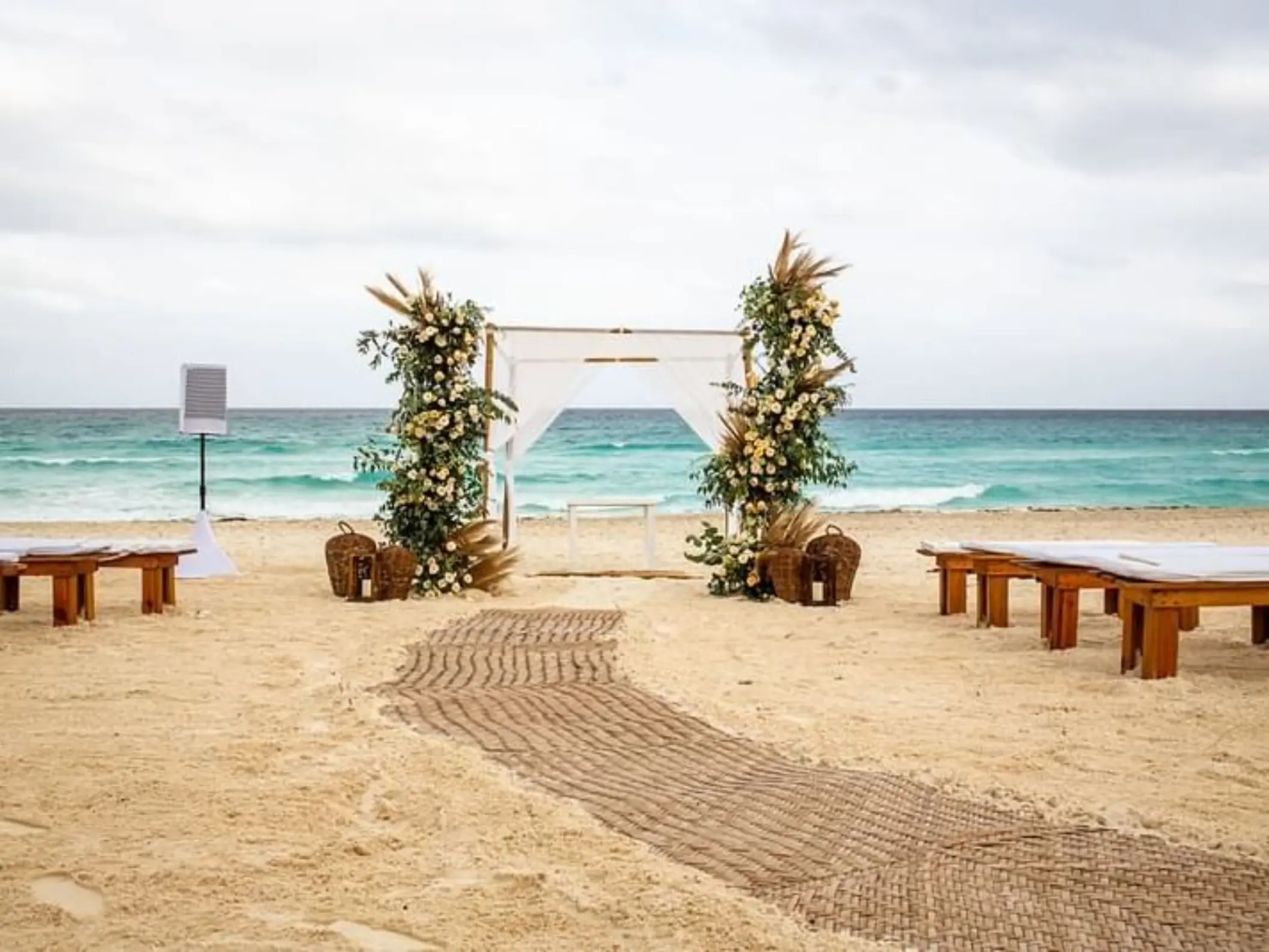 A wedding venue on the beach with a white gazebo, floral arrangements, and benches on the sand.