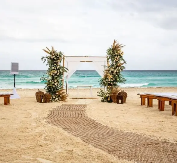 A wedding venue on the beach with a white gazebo, floral arrangements, and benches on the sand.
