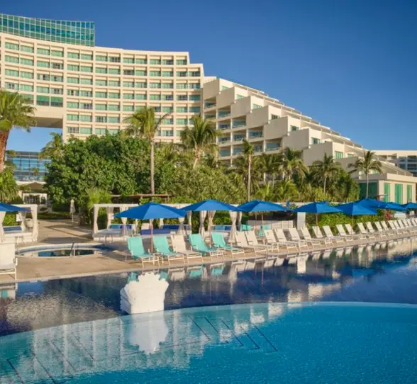 A resort pool area with lounge chairs and umbrellas, with the resort building and palm trees in the background.