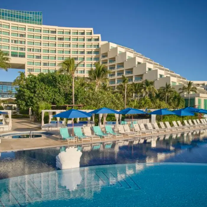 A resort pool area with lounge chairs and umbrellas, with the resort building and palm trees in the background.