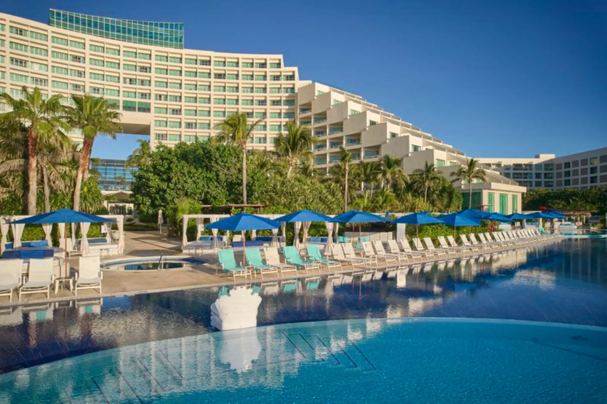 A resort pool area with lounge chairs and umbrellas, with the resort building and palm trees in the background.