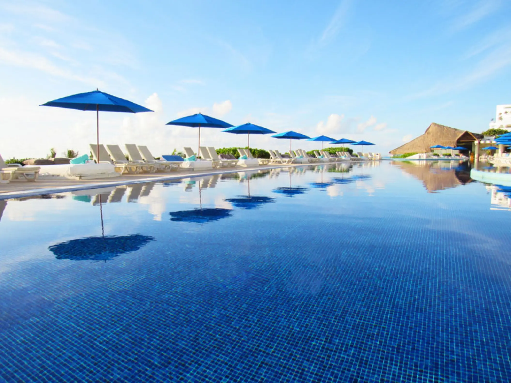 A long infinity pool with blue tiles, reflecting lounge chairs, and blue umbrellas against a clear sky.