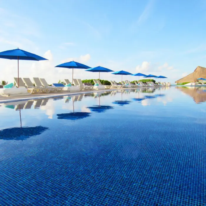A long infinity pool with blue tiles, reflecting lounge chairs, and blue umbrellas against a clear sky.