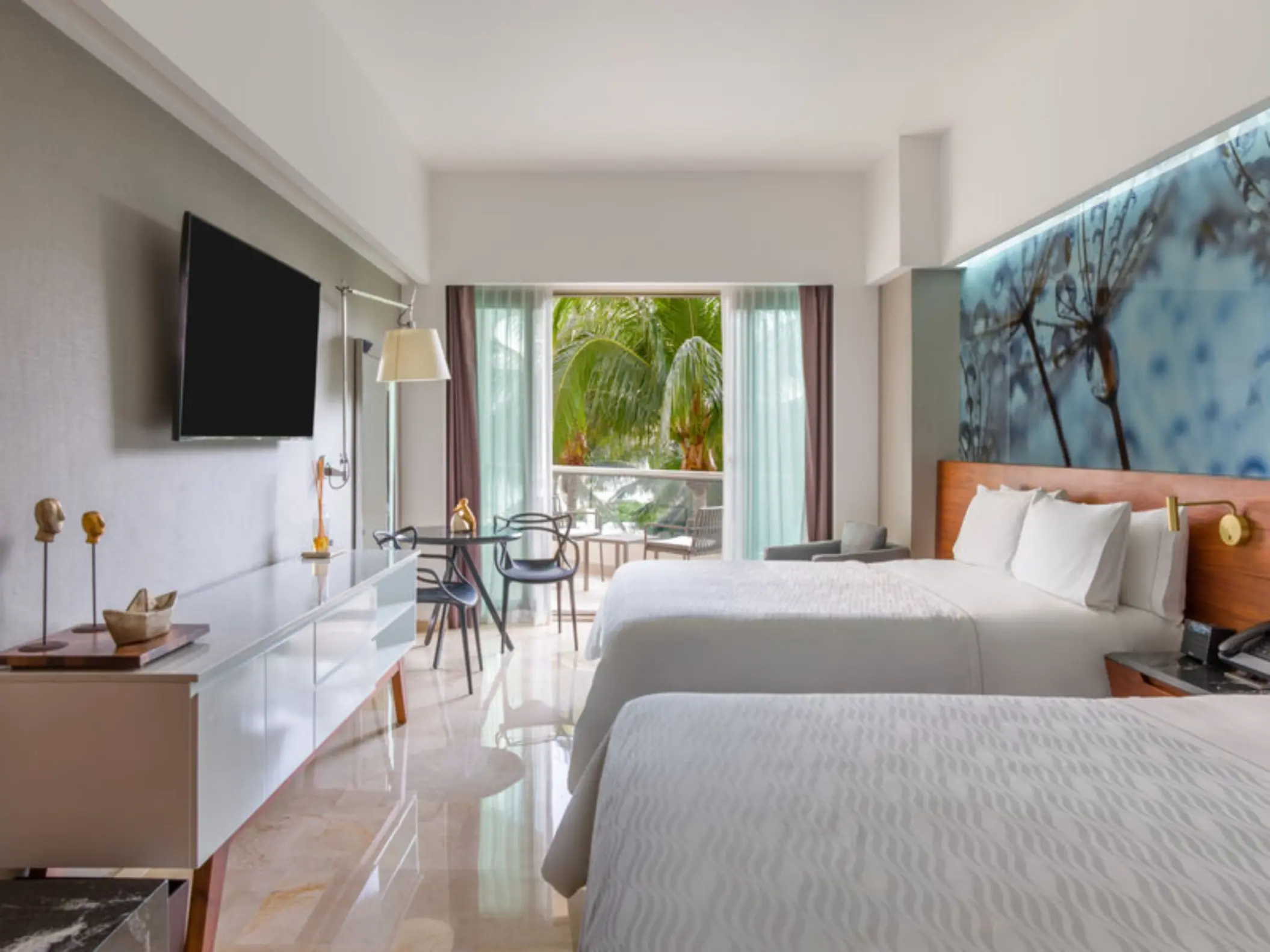 A hotel bedroom with two beds, a small dining table, and a balcony with a view of a garden.
