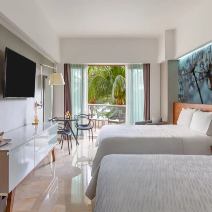 A hotel bedroom with two beds, a small dining table, and a balcony with a view of a garden.
