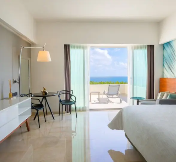 A hotel room with a large bed, a television, and a balcony overlooking a bright blue ocean.