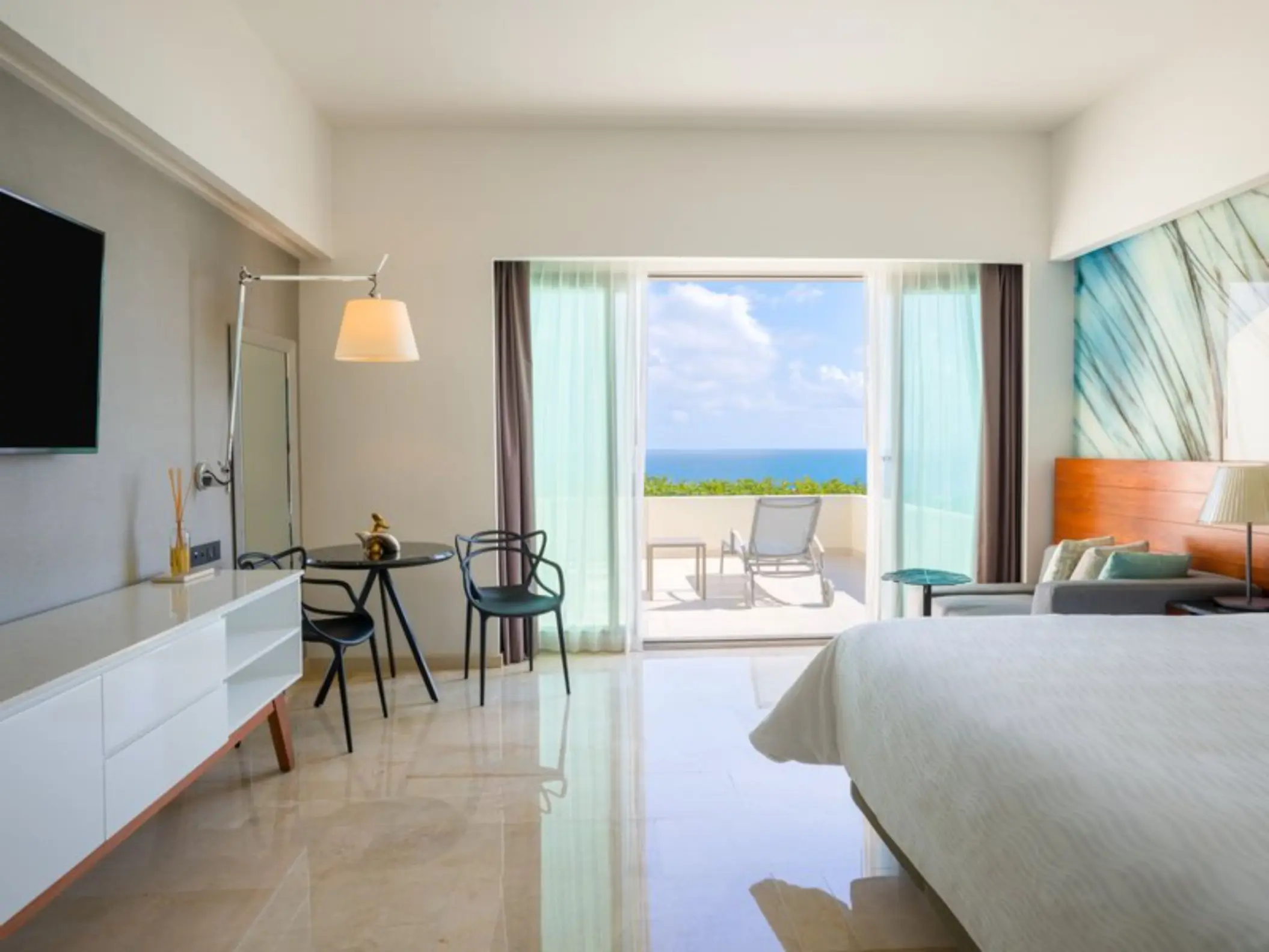 A hotel room with a large bed, a television, and a balcony overlooking a bright blue ocean.
