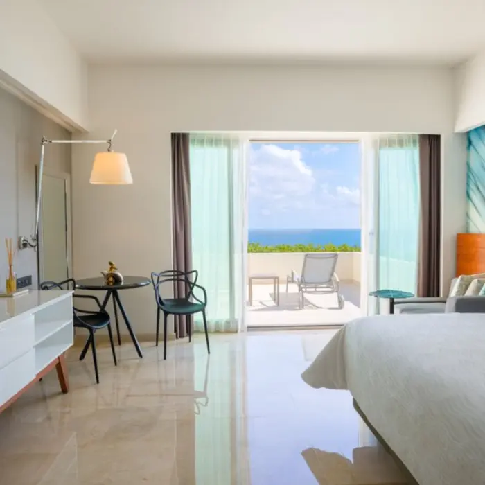 A hotel room with a large bed, a television, and a balcony overlooking a bright blue ocean.