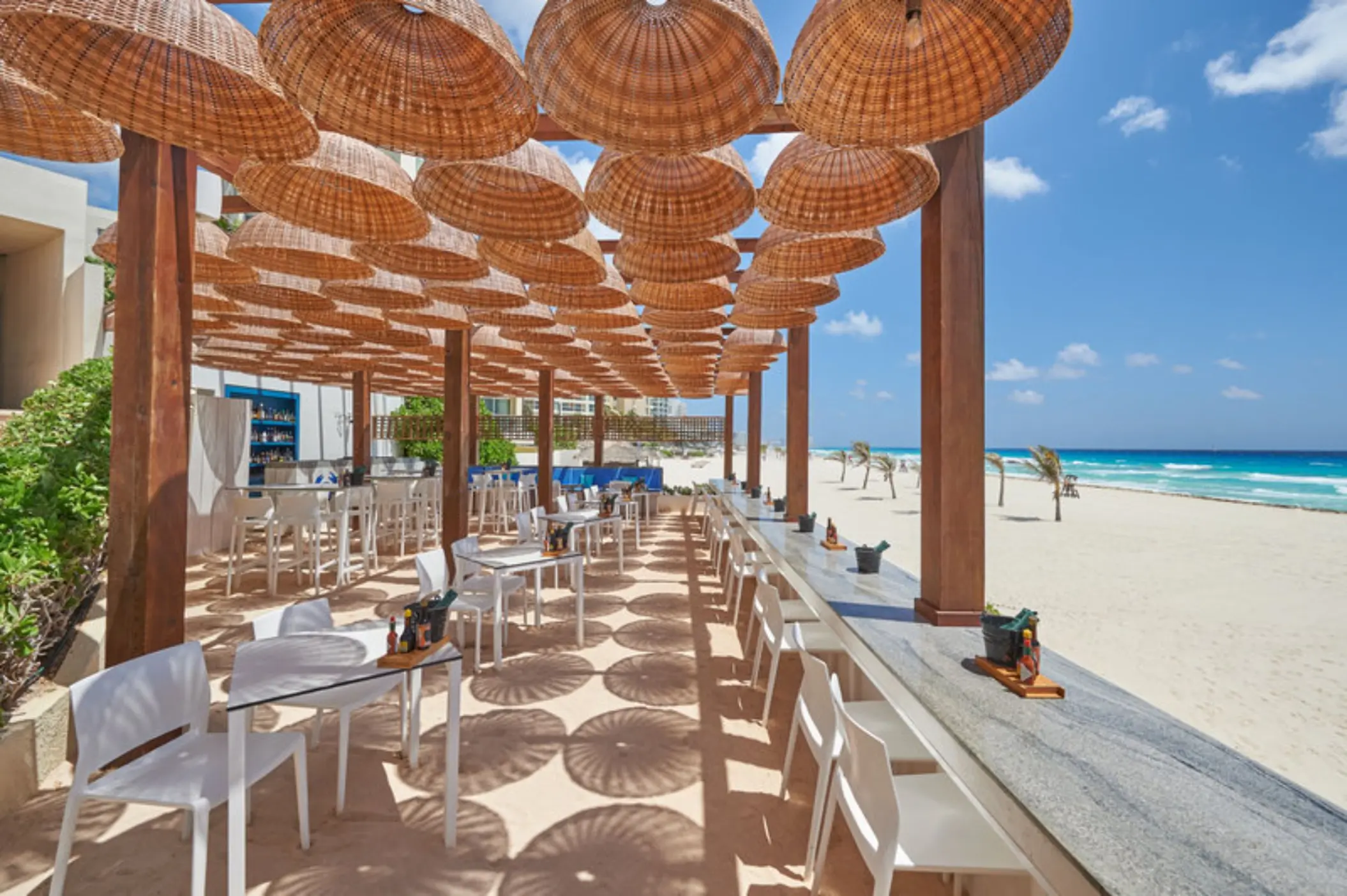 An outdoor beach bar and restaurant with white tables, chairs, and a thatched-roof cover.