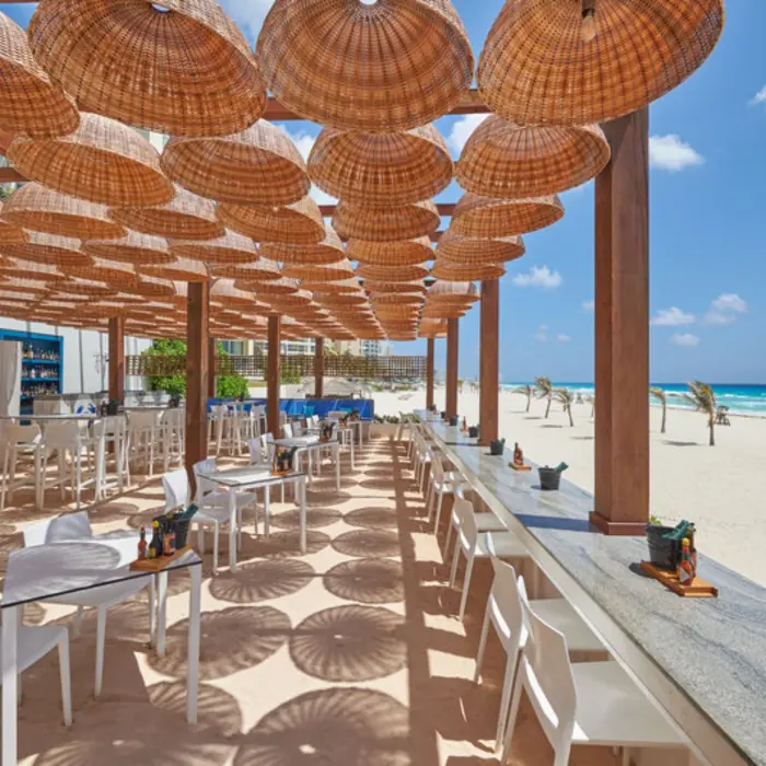 An outdoor beach bar and restaurant with white tables, chairs, and a thatched-roof cover.