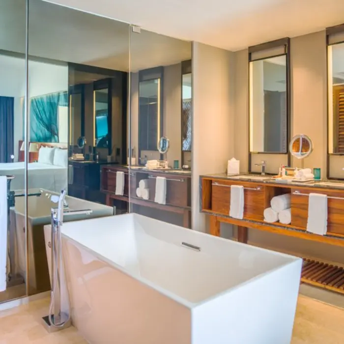 A clean, modern bathroom with a freestanding white tub and a large glass window leading to the bedroom.