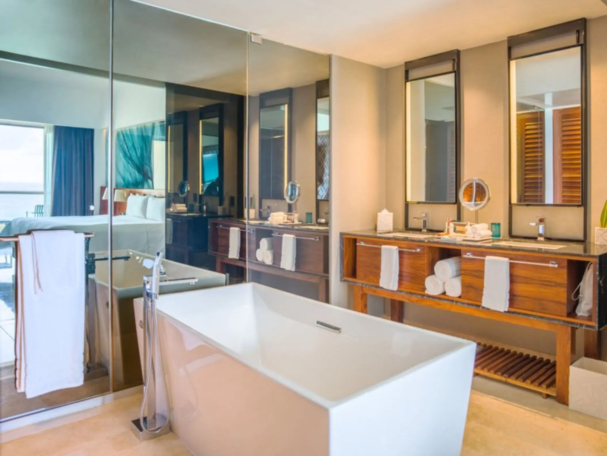A clean, modern bathroom with a freestanding white tub and a large glass window leading to the bedroom.