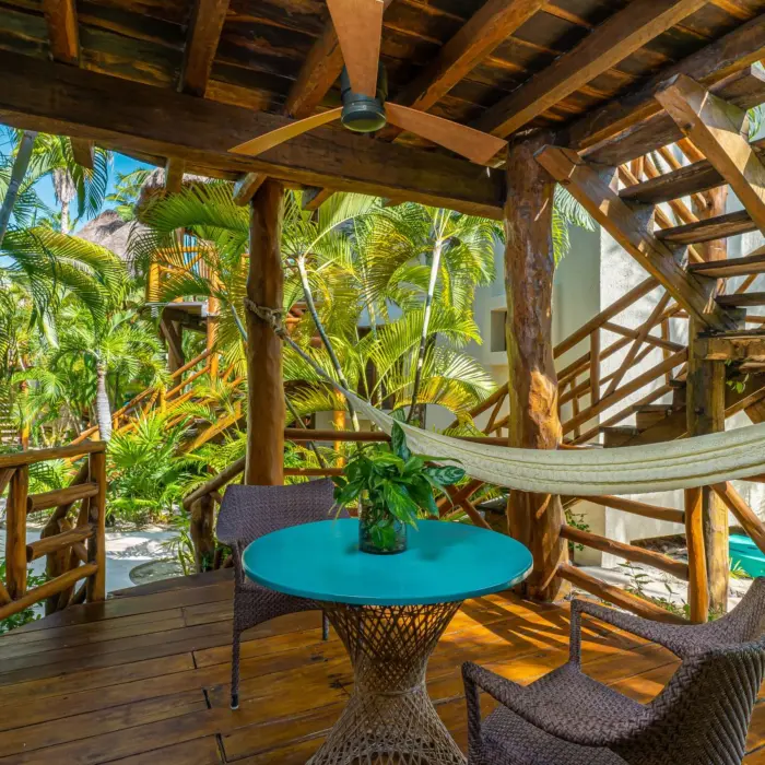 Shaded terrace with a hammock and turquoise table nestled in dense jungle greenery and wooden stairs.
