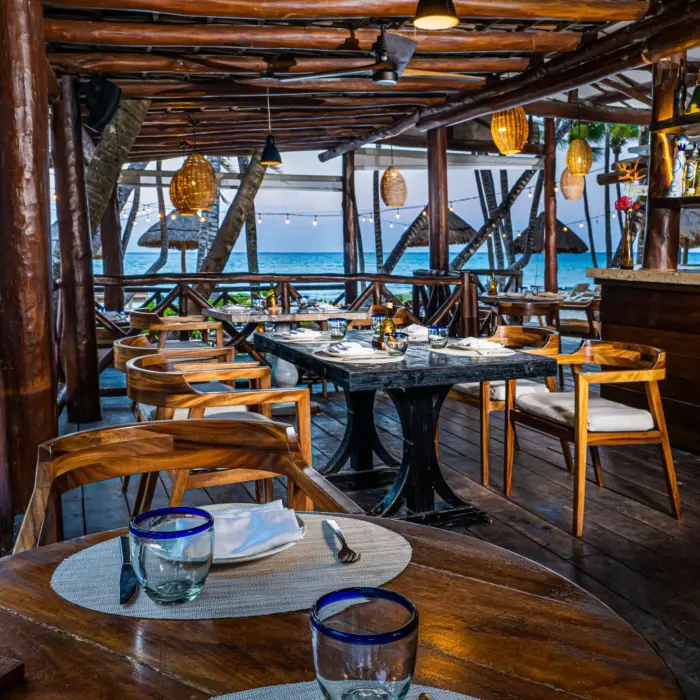 Seaside open-air restaurant with wooden furniture, bar, and string lights over the ocean.
