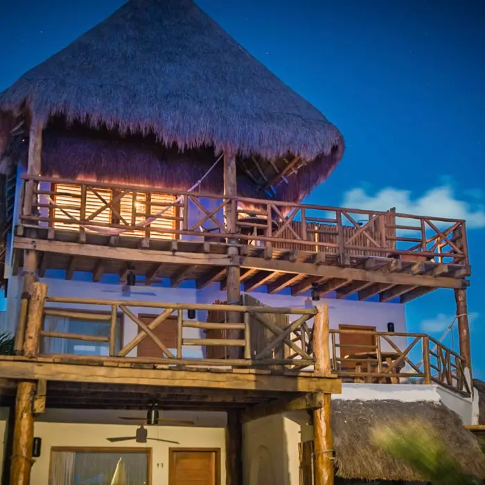 Rustic three-level beachfront villa with thatched roof, wood balconies, and warm night lighting by the sea.