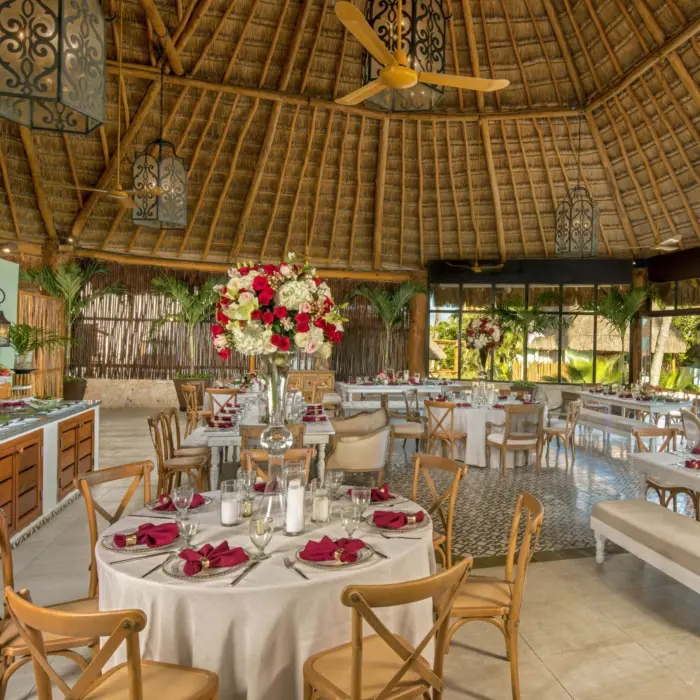 Indoor wedding reception with round tables, floral centerpieces, and a thatched roof at a beachside resort.