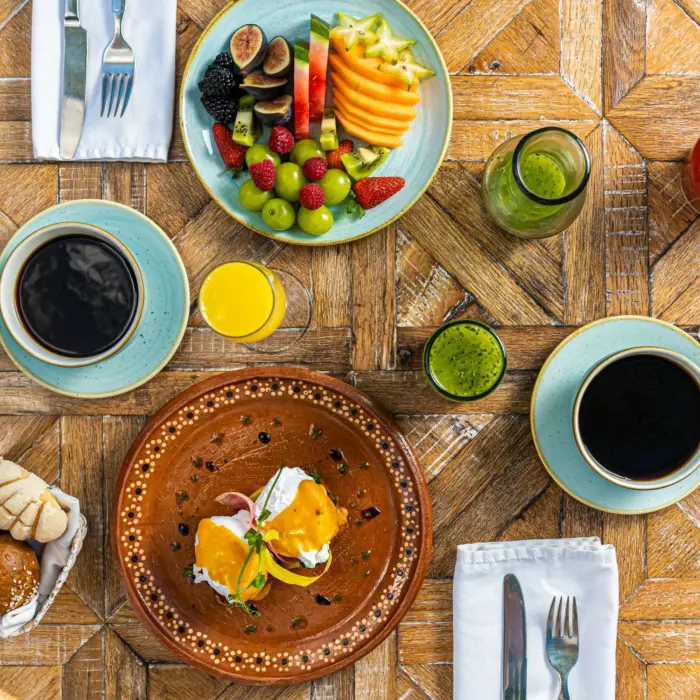 Colorful breakfast spread with tropical fruit, fresh pastries, eggs benedict, and green juice on a rustic wood table.