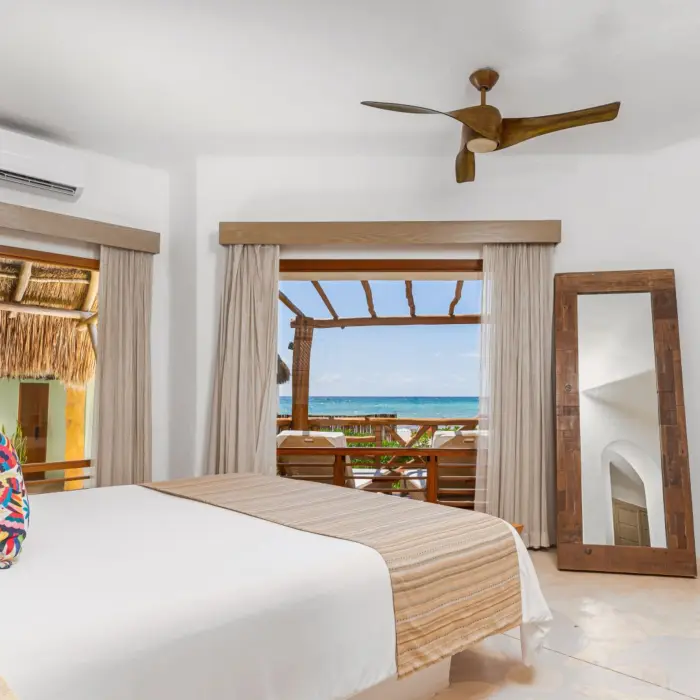 Oceanfront bungalow bedroom with ceiling fan, patio view, and rustic design featuring palapa roof and wood accents.