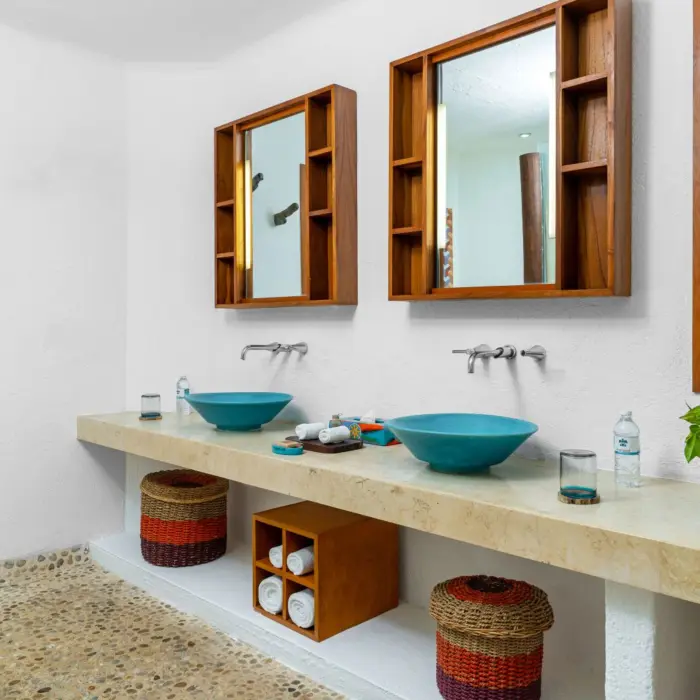Spacious bathroom with dual turquoise sinks, wood mirrors, pebble floor, and tropical greenery visible through the windows.