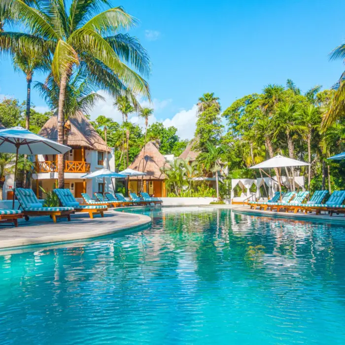 Large resort pool surrounded by lush palm trees, loungers, and thatched-roof suites in the background.