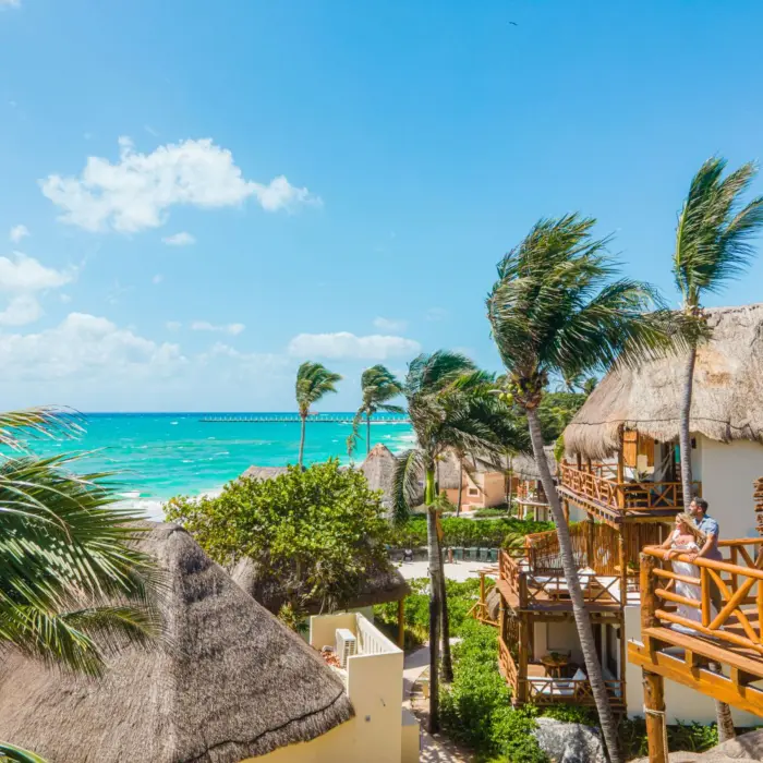 Couple enjoying the sea breeze on a private oceanview balcony surrounded by thatched-roof bungalows and palm trees.