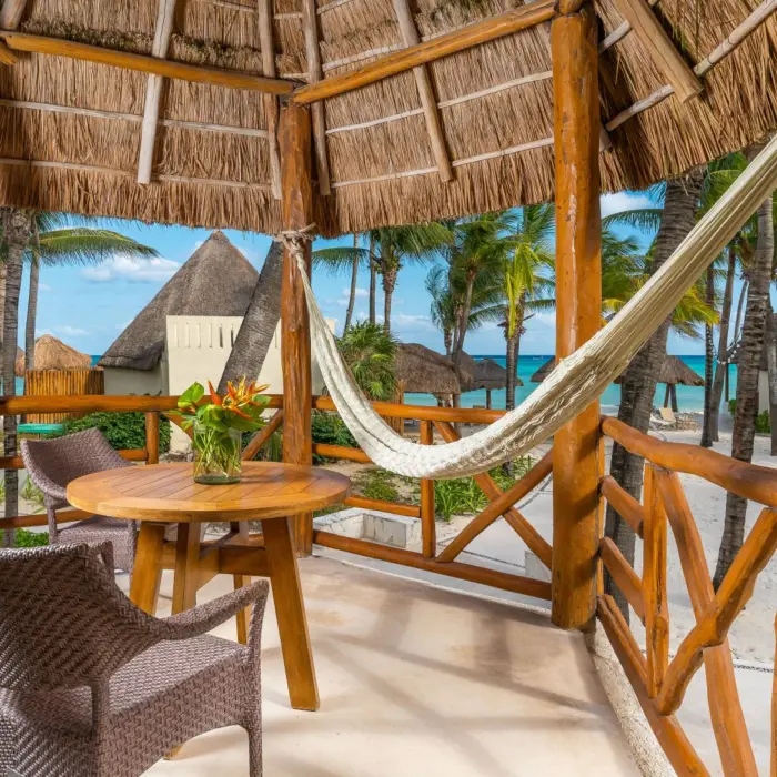 Thatched-roof terrace with hammock, tropical flowers, and a wooden table overlooking palm trees and the beach.