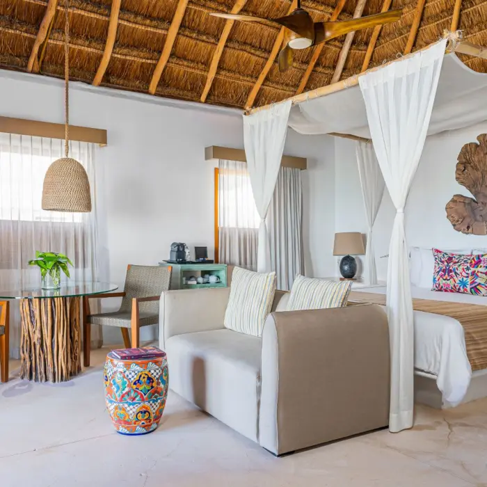 Spacious suite with canopy bed, sitting area, dining table, handmade décor, and palapa roof overhead.