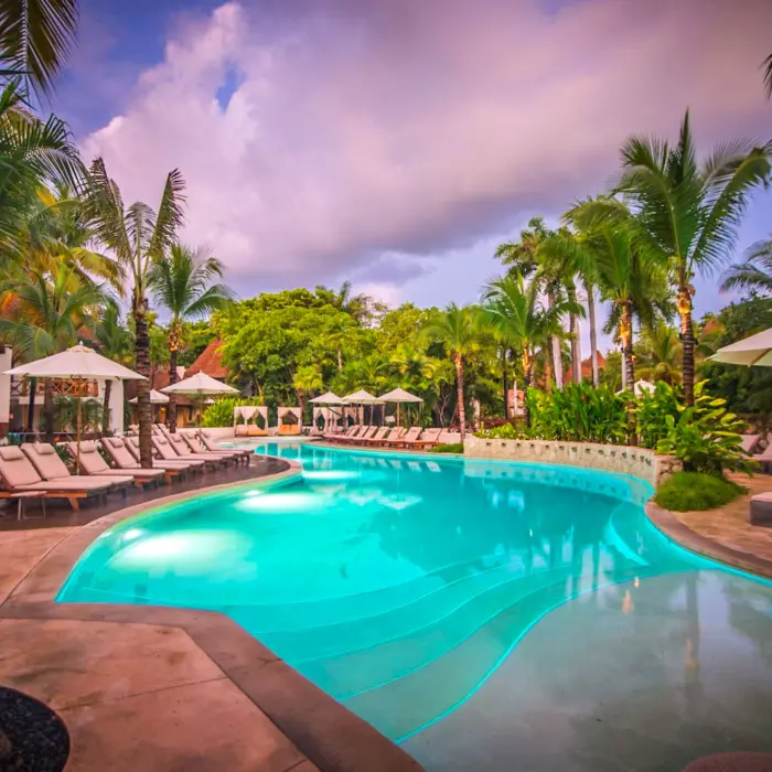 Resort swimming pool at twilight surrounded by sun loungers, umbrellas, and dense tropical gardens.