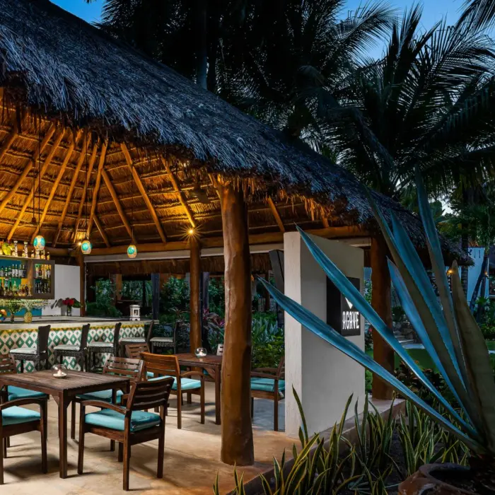 Open-air Agave bar under a palapa with mosaic tile details, turquoise seating, and glowing evening lights.