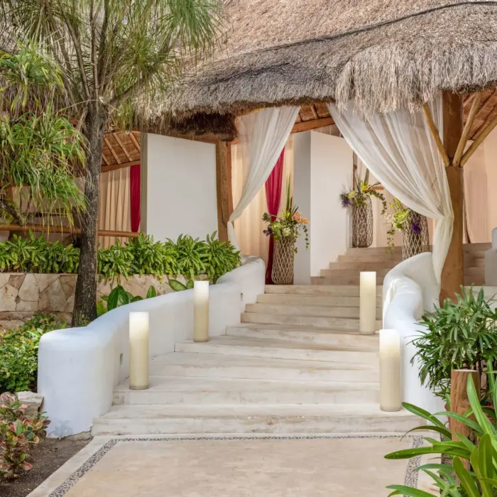 Elegant entrance with curved staircase, tall candles, and white drapery framed by tropical plants and palms.