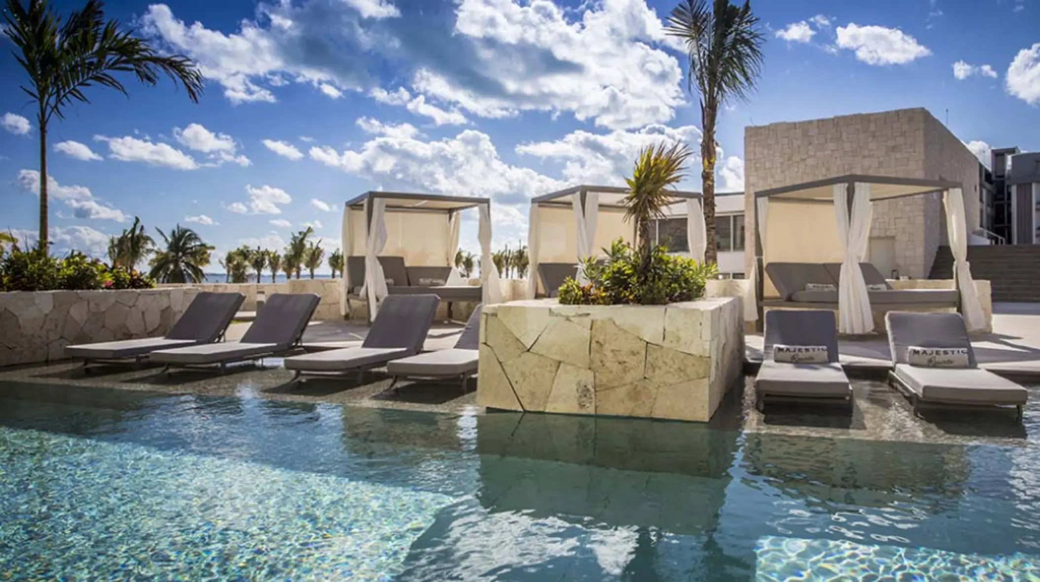 Poolside cabanas and loungers with canopy curtains at Majestic Elegance Costa Mujeres under sunny skies.