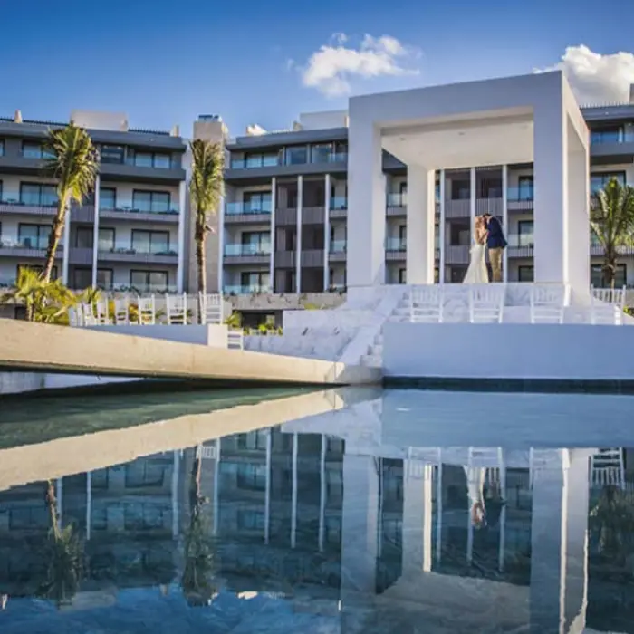 White outdoor wedding gazebo with water reflection, palm trees, and resort buildings in the background.