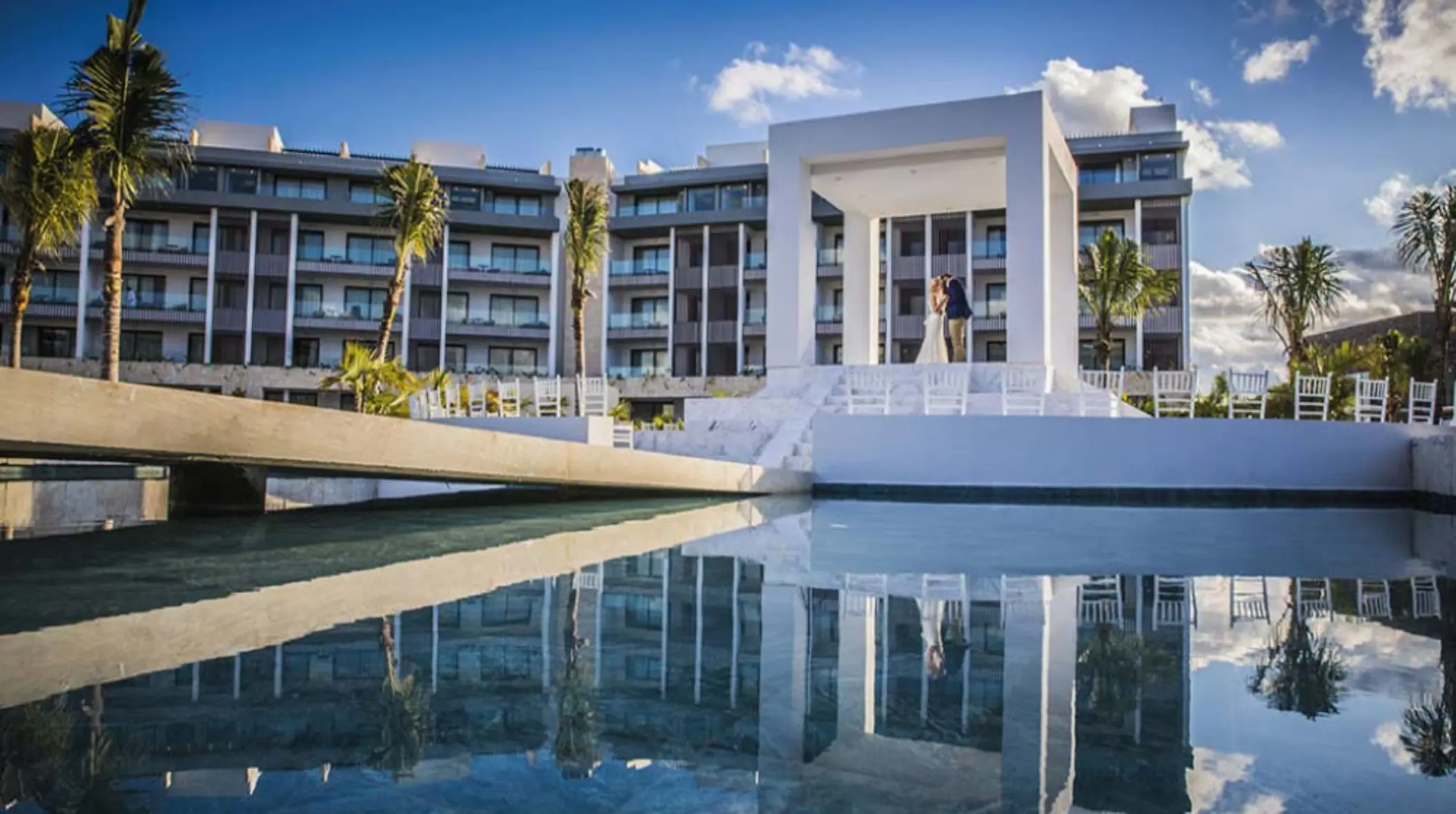 White outdoor wedding gazebo with water reflection, palm trees, and resort buildings in the background.