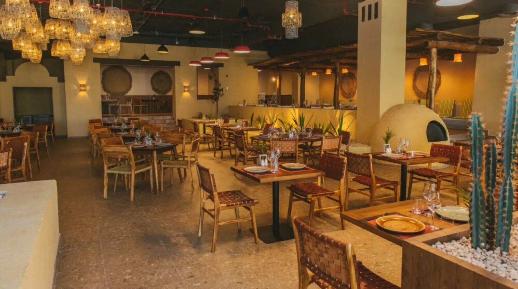 Rustic Mexican restaurant with earthy tones, cactus decor, woven chairs, and clay oven centerpiece.