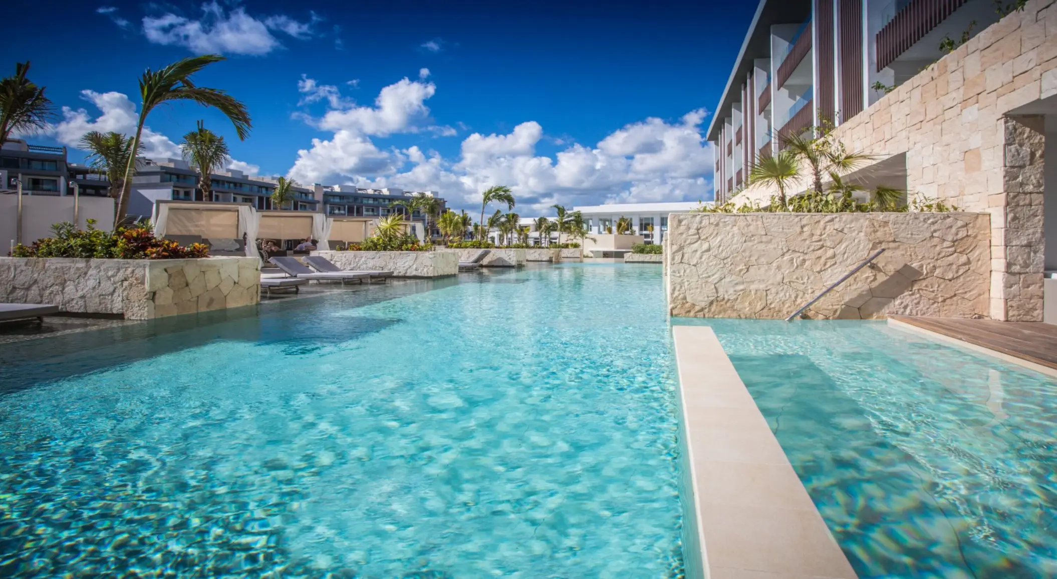 Expansive outdoor pool with palm trees, stone walls, and resort buildings under bright blue skies.