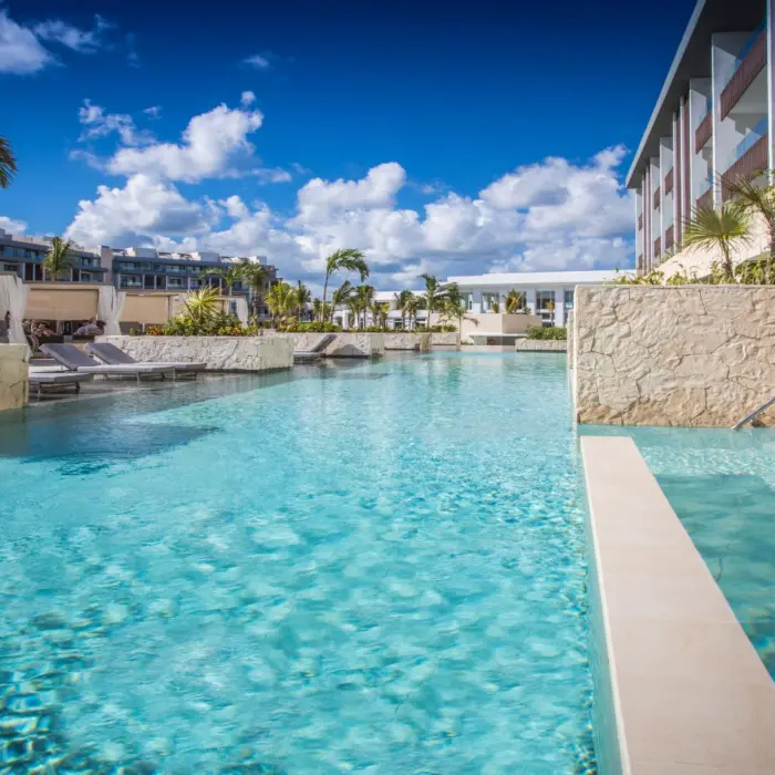 Expansive outdoor pool with palm trees, stone walls, and resort buildings under bright blue skies.