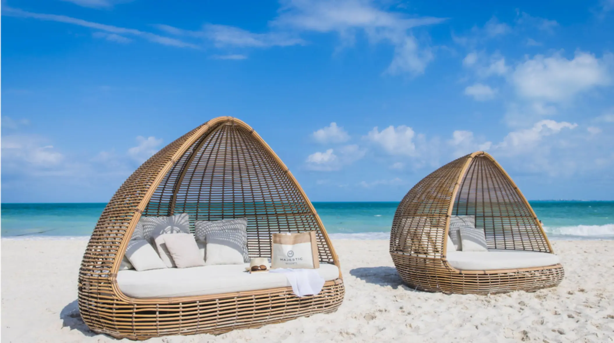 Stylish beach cabanas on white sand, shaded by wicker domes with ocean views and Majestic-branded accessories.