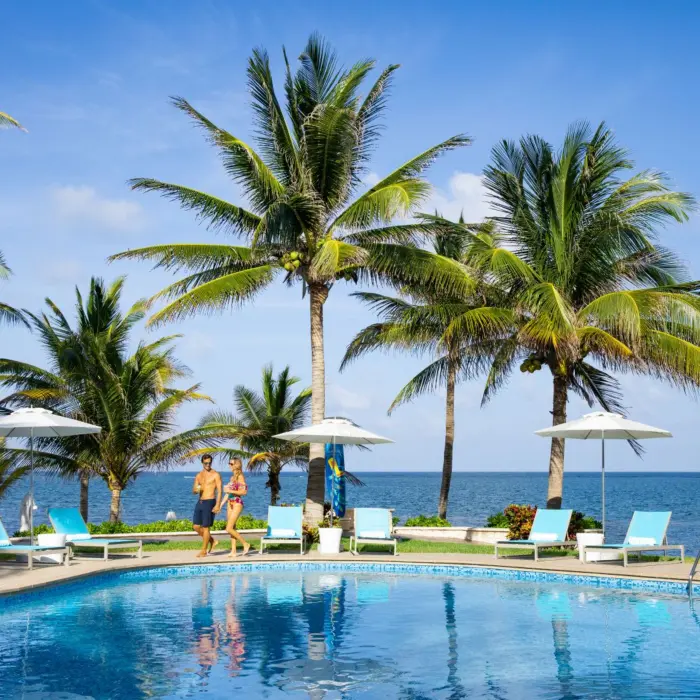 Couple walking by a tropical pool with lounge chairs and umbrellas surrounded by palm trees near the ocean.