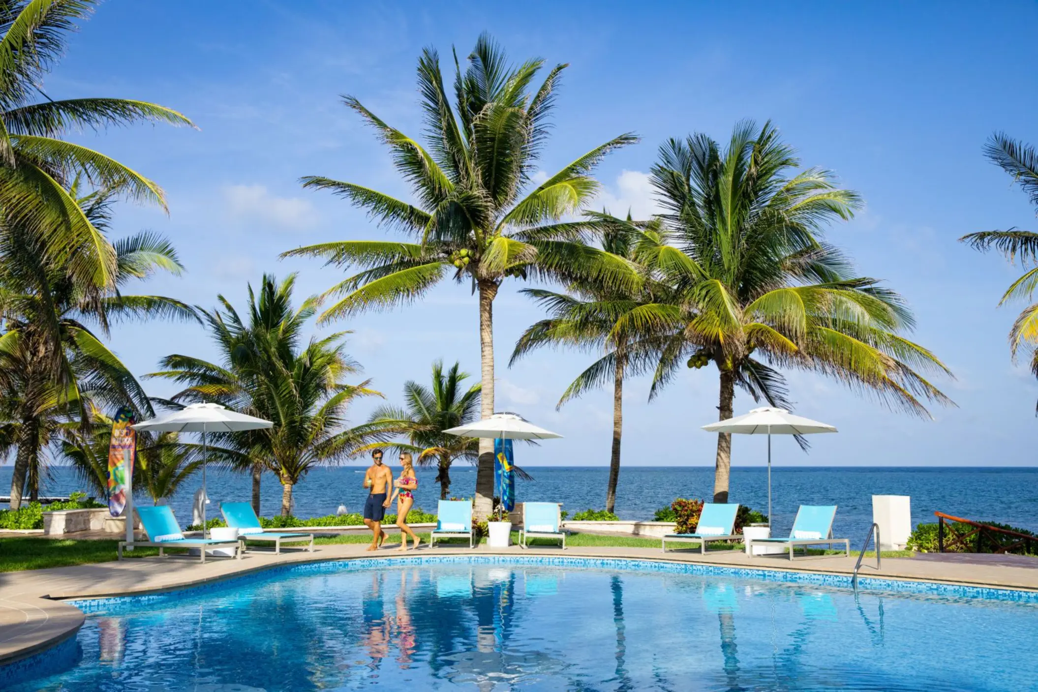 Couple walking by a tropical pool with lounge chairs and umbrellas surrounded by palm trees near the ocean.
