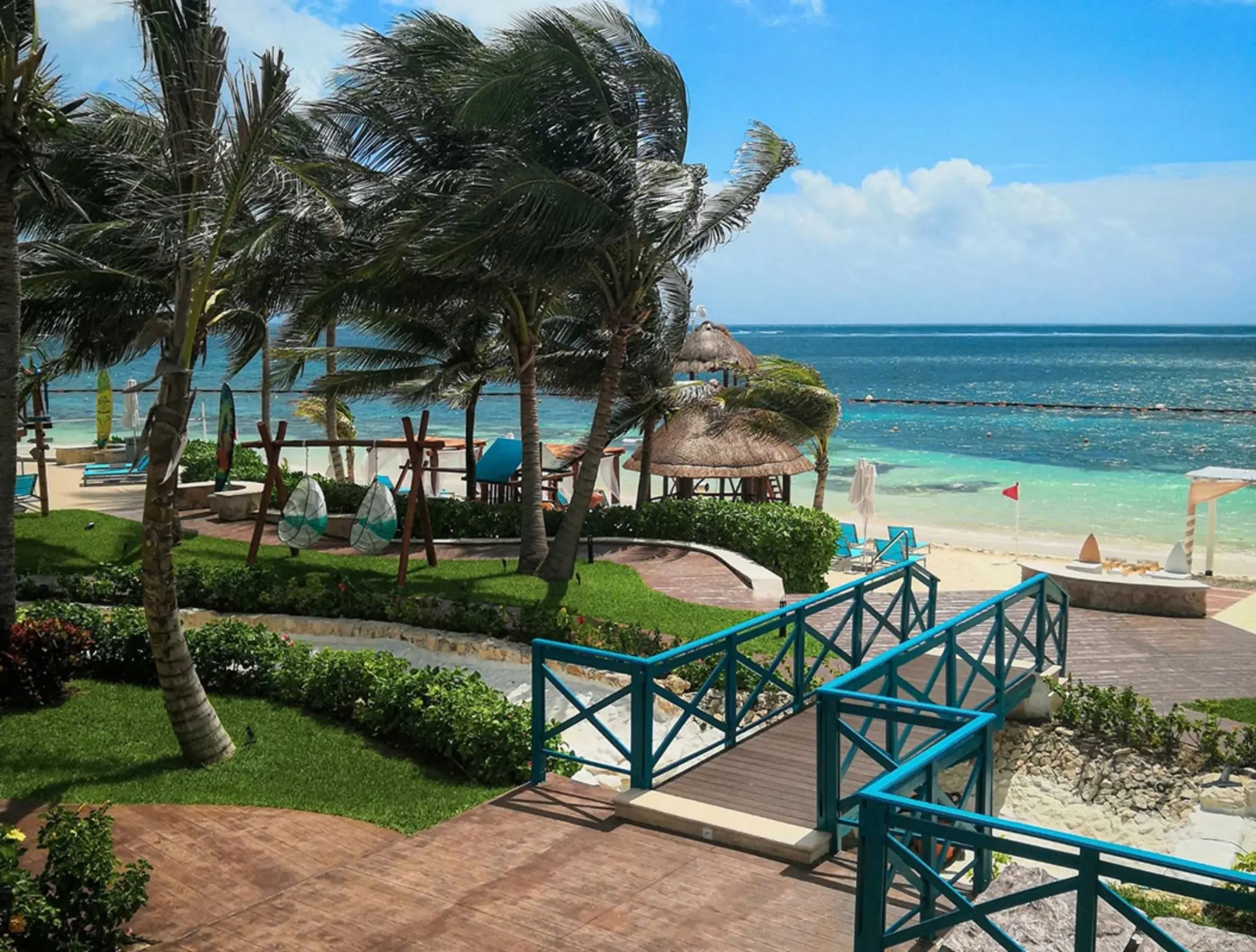 Oceanfront view with turquoise fences, palm trees, and cabanas overlooking a clear Caribbean sea.