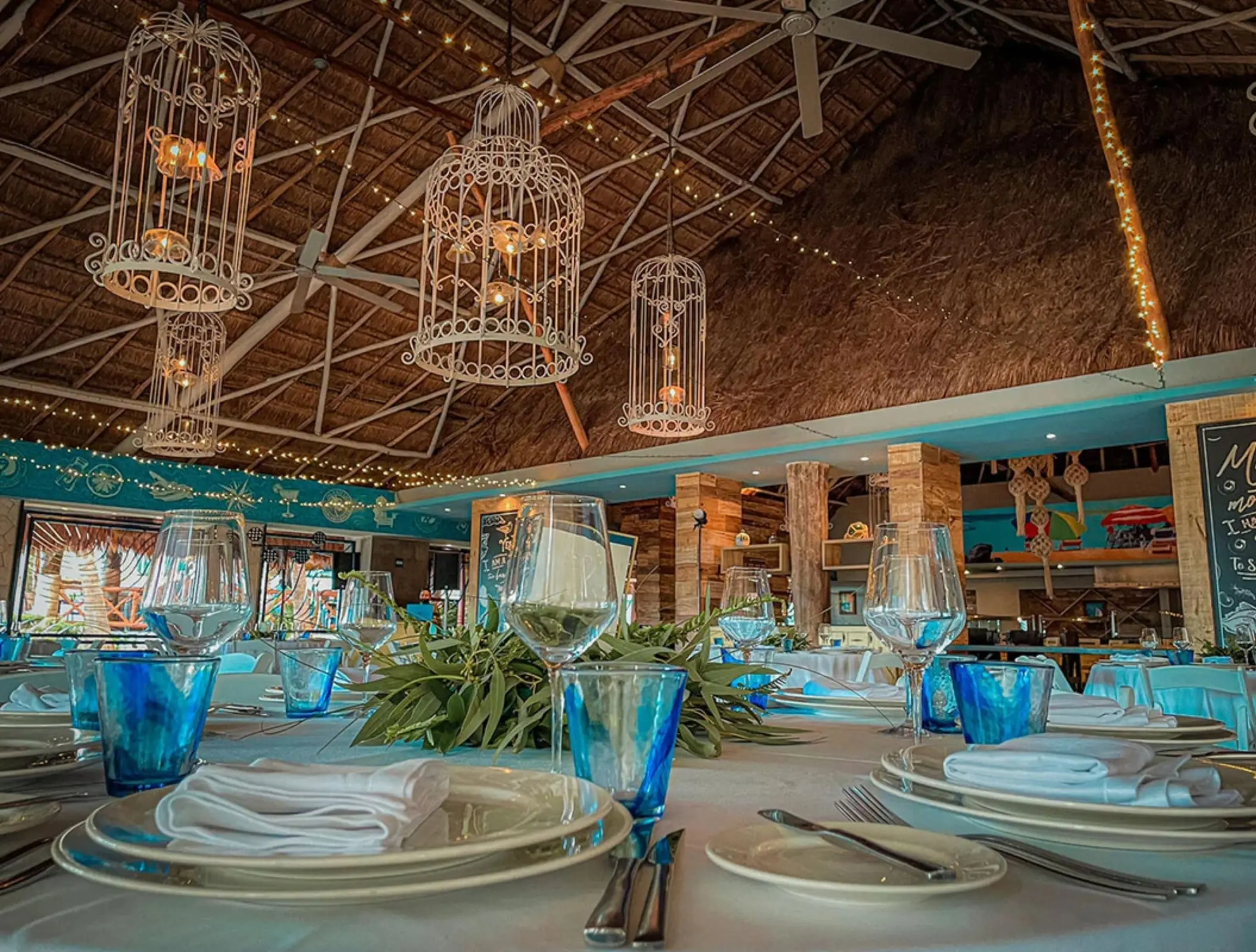 Close-up of a rustic-chic restaurant setup with blue accents, wine glasses, and twinkle lights under a palapa.