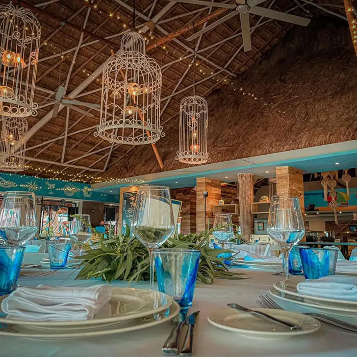 Close-up of a rustic-chic restaurant setup with blue accents, wine glasses, and twinkle lights under a palapa.