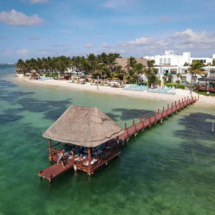 A thatched-roof pier pavilion extends over turquoise water in front of a tropical beachfront resort.