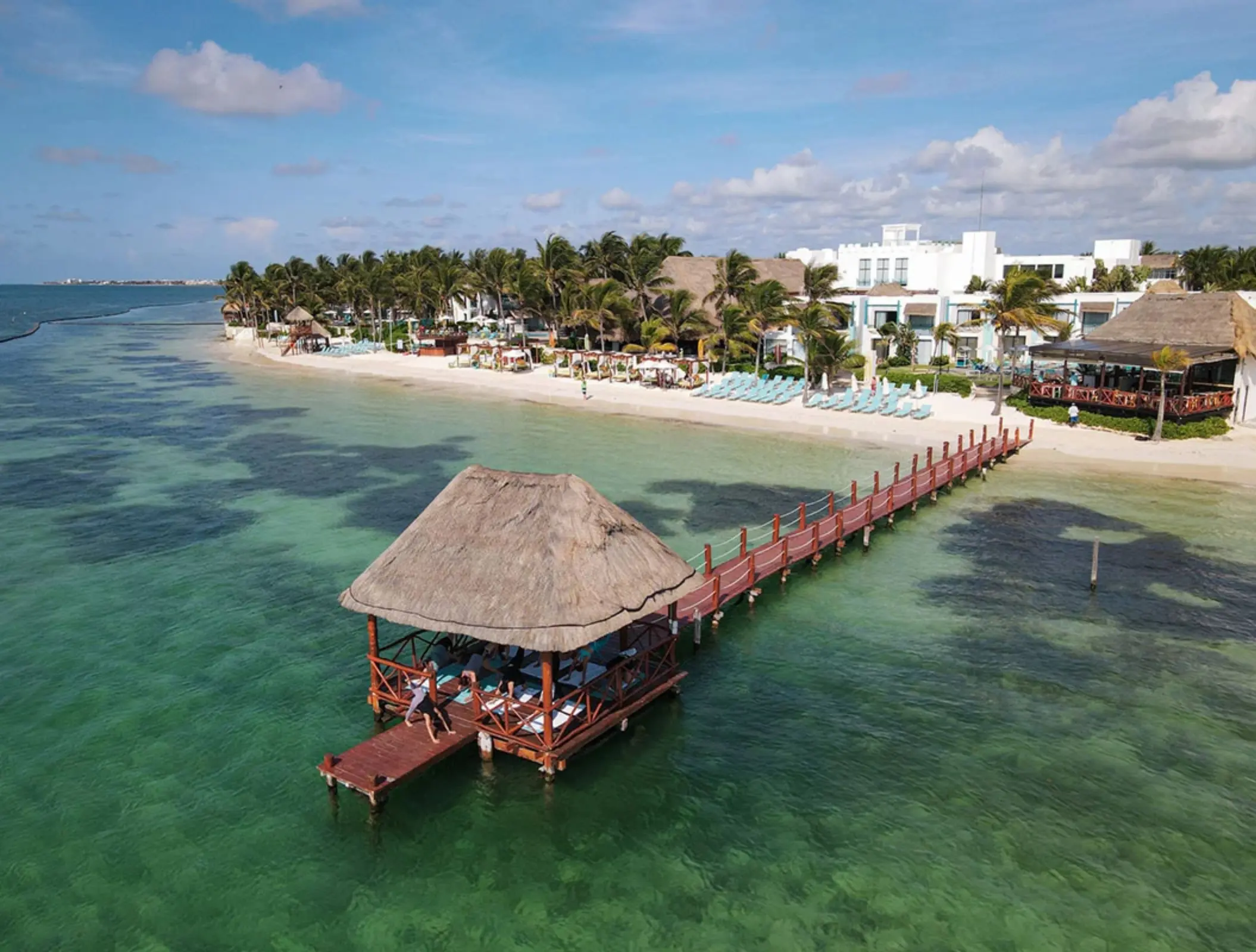 A thatched-roof pier pavilion extends over turquoise water in front of a tropical beachfront resort.