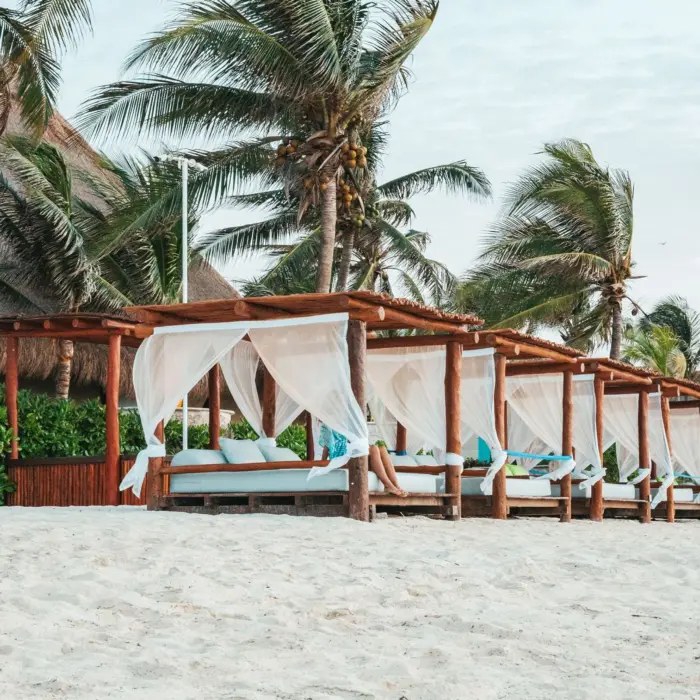 Shaded beach cabanas with white curtains and blue cushions surrounded by palm trees and soft white sand.