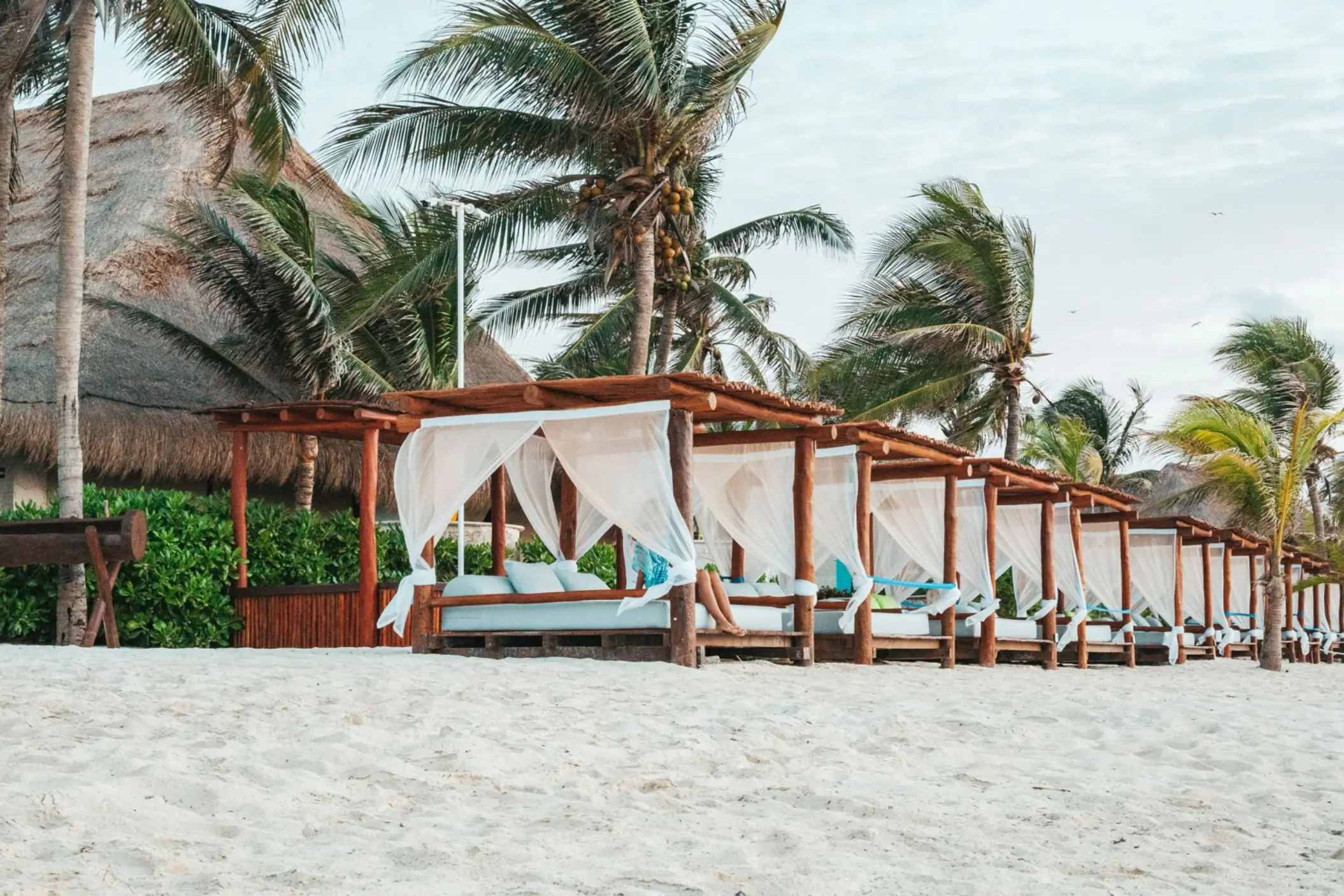 Shaded beach cabanas with white curtains and blue cushions surrounded by palm trees and soft white sand.