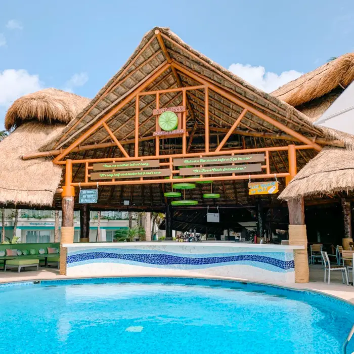 Swim-up palapa bar with tropical decor, poolside seating, and colorful signs under a thatched roof by the pool.