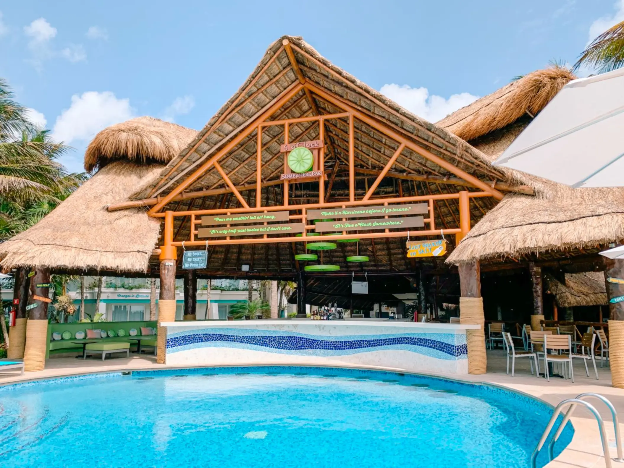 Swim-up palapa bar with tropical decor, poolside seating, and colorful signs under a thatched roof by the pool.